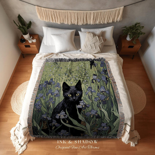 May include: A woven throw blanket featuring a black cat amidst blue irises and green foliage. The blanket has a fringed edge and the text "INK & SHADOW Original Fine Art Throws" at the bottom.