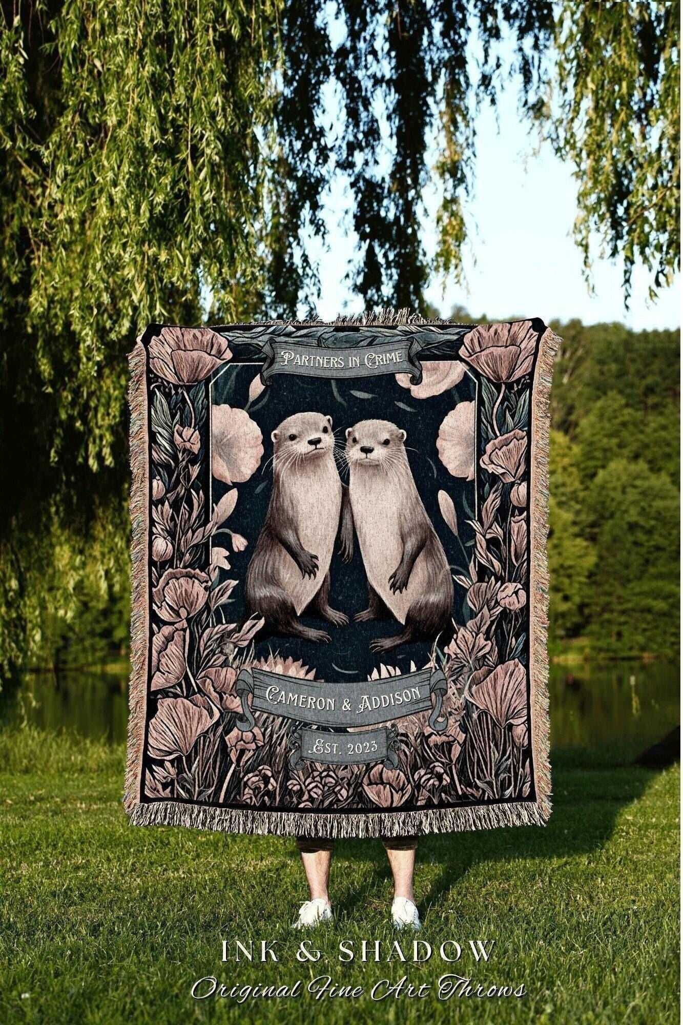 May include: A decorative throw blanket featuring two otters, surrounded by floral designs, with the text "PARTNERS IN CRIME" and "CAMERON & ADDISON Est. 2023". The blanket has a dark background with a fringed edge. The words "INK & SHADOW Original Fine Art Throws" are at the bottom.