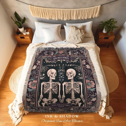 May include: A dark green throw blanket with a design featuring two skeletons, flowers, and the words "Kindred Spirits" and "Dylan & Gabby 2024". The blanket has a fringed edge and is displayed on a bed.