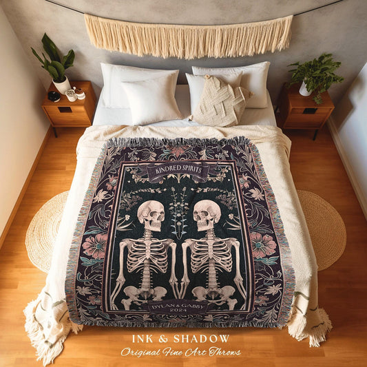 May include: A dark green throw blanket with a design featuring two skeletons, flowers, and the words "Kindred Spirits" and "Dylan & Gabby 2024". The blanket has a fringed edge and is displayed on a bed.