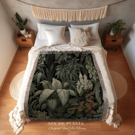May include: A woven throw blanket with a botanical print of various green plants and foliage. The blanket is draped over a bed with white bedding. The text "JOY OF PLANTS Original Fine Art Throws" is printed on the bottom.