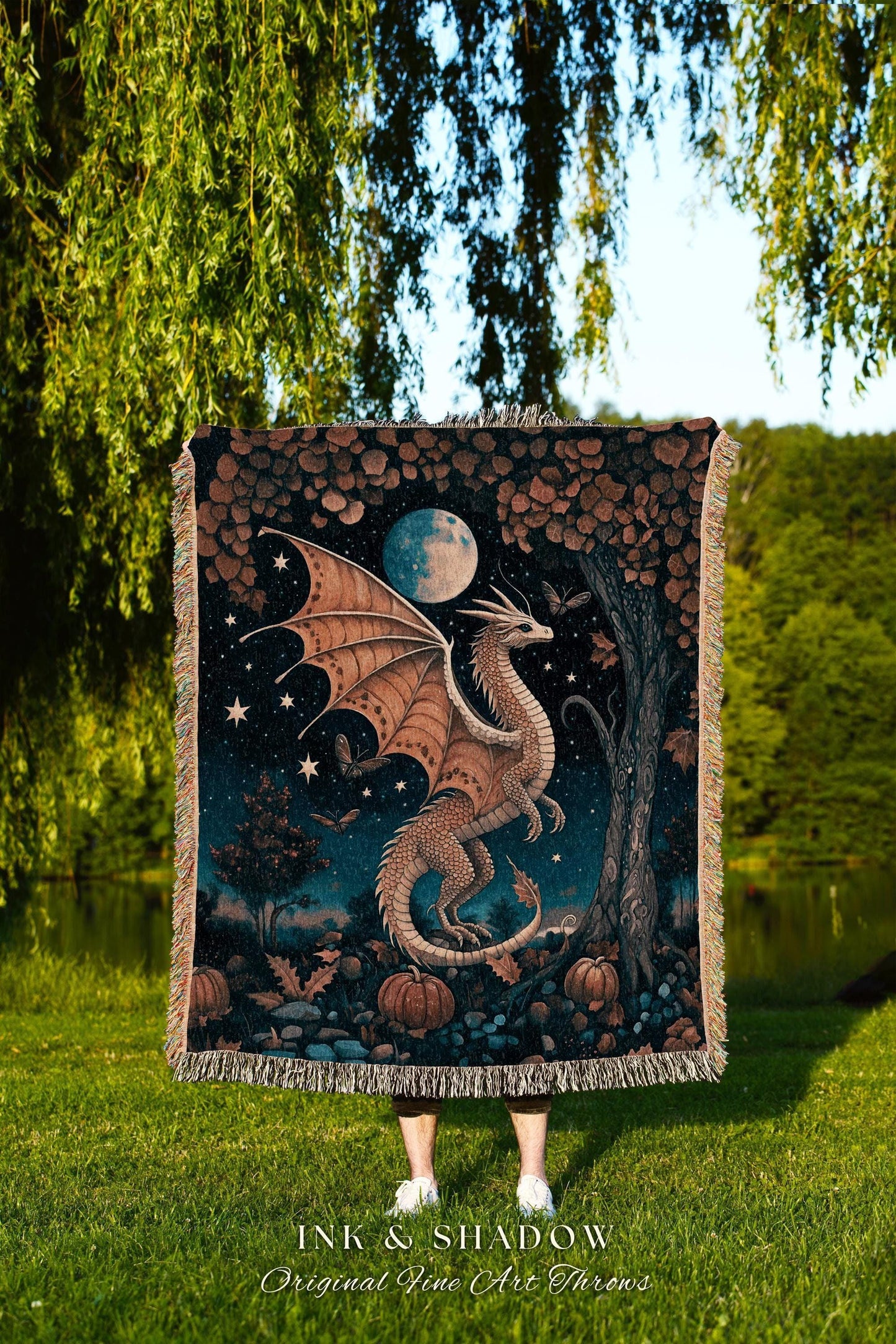 May include: A woven throw blanket featuring a dragon design. The dragon is depicted with tan wings and a white body, set against a night sky with a full moon, stars, and pumpkins. The blanket has a fringed edge.
