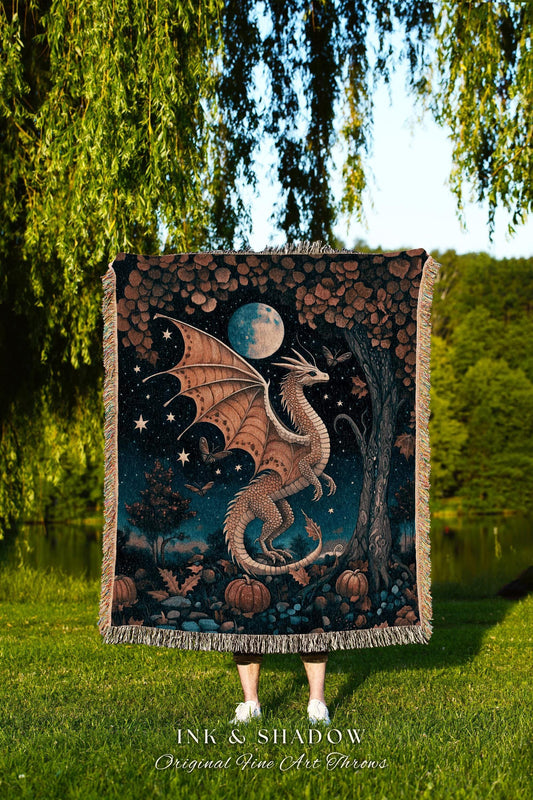 May include: A woven throw blanket featuring a dragon design. The dragon is depicted with tan wings and a white body, set against a night sky with a full moon, stars, and pumpkins. The blanket has a fringed edge.