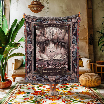 May include: A tapestry throw blanket featuring a romantic scene with two swans forming a heart shape on a lake. The blanket has a dark color palette with floral and foliage accents, and the text "THE LOVERS" is displayed at the top.