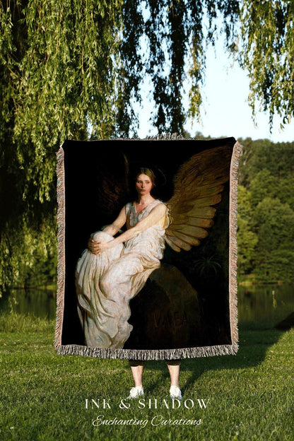 May include: A black throw blanket featuring a classical painting of a seated angel with large wings. The angel is wearing a white dress and is seated on a rock. The blanket has a fringed edge and the text "INK & SHADOW Enchanting Curations" is at the bottom.