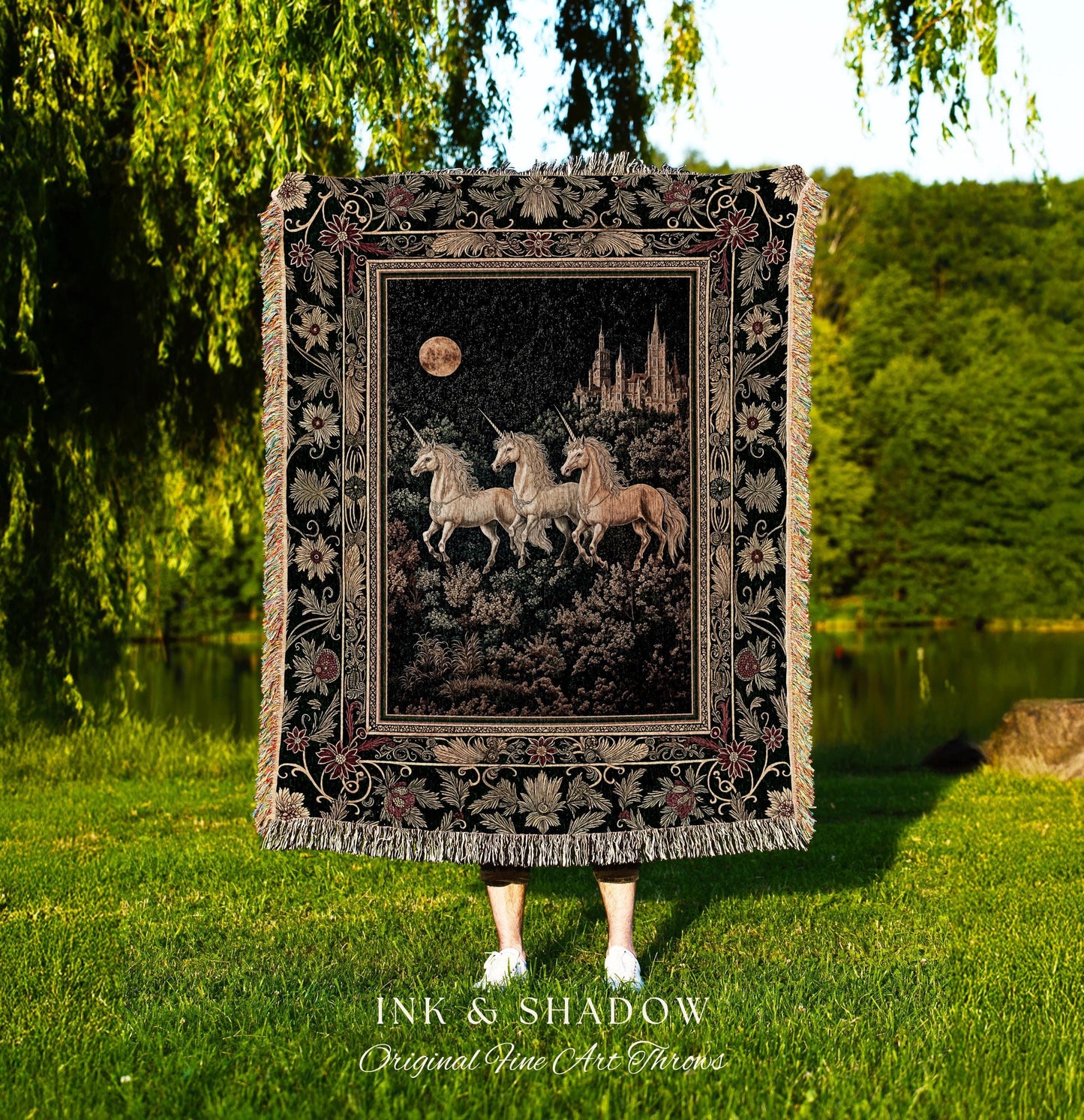 May include: A tapestry throw blanket featuring a central image of three unicorns in a forest under a full moon, with a castle in the background. The blanket has a black background with floral and leaf border designs and fringe.