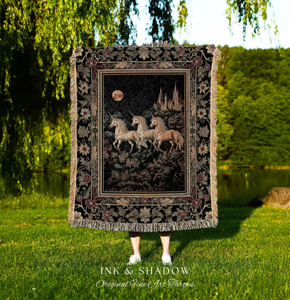 May include: A tapestry throw blanket featuring a central image of three unicorns in a forest under a full moon, with a castle in the background. The blanket has a black background with floral and leaf border designs and fringe.