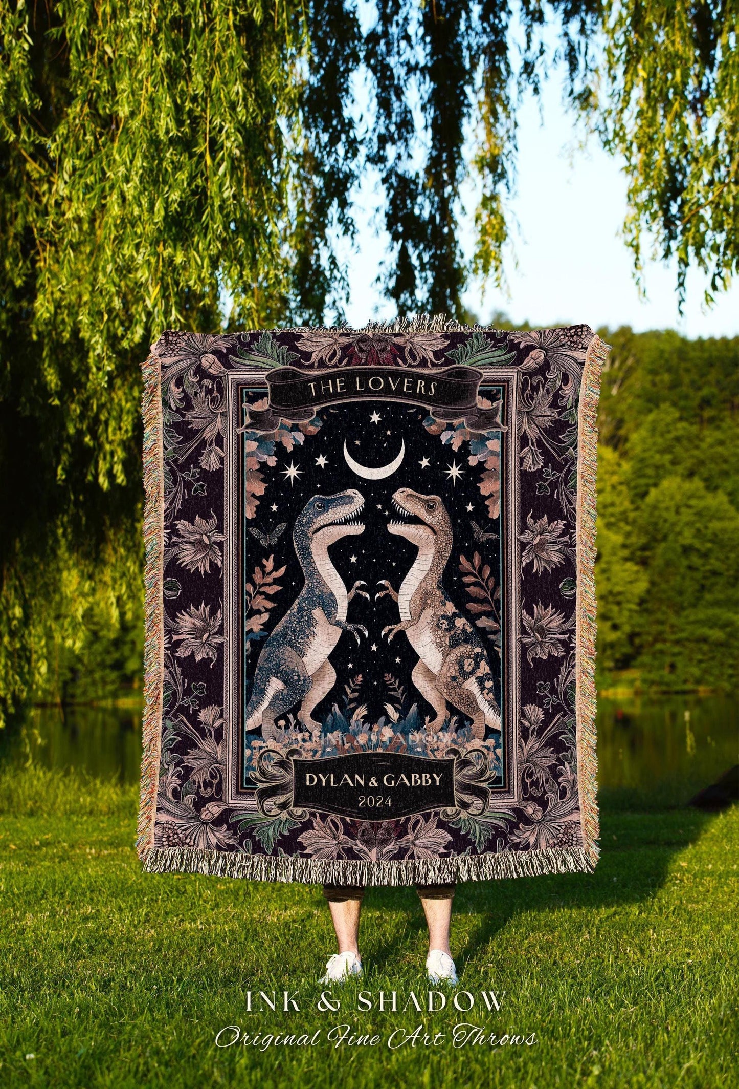 May include: A woven throw blanket featuring a dark background with two dinosaur illustrations facing each other. The blanket has a decorative border, the words "THE LOVERS", and the names "DYLAN & GABBY" with the year 2024.