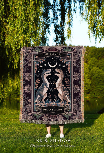 May include: A woven throw blanket featuring a dark background with two dinosaur illustrations facing each other. The blanket has a decorative border, the words "THE LOVERS", and the names "DYLAN & GABBY" with the year 2024.