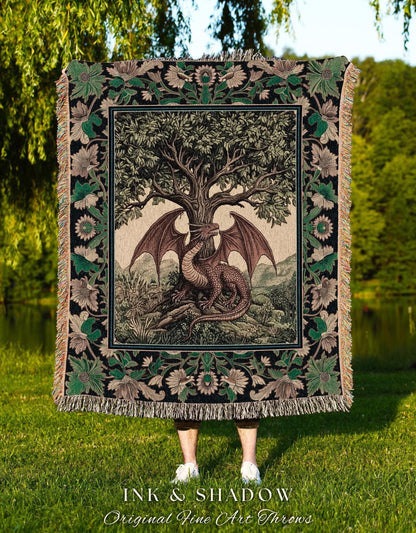 May include: A tapestry throw blanket featuring a detailed illustration of a dragon resting beneath a large tree. The dragon is brown with large wings. The blanket has a black border with floral and leaf patterns and fringe.