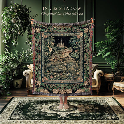 May include: A tapestry throw blanket featuring a large frog on a rock with a castle in the background. The throw has a dark green and black floral border and fringe. The text "INK & SHADOW Original Fine Art Throws" is at the top.