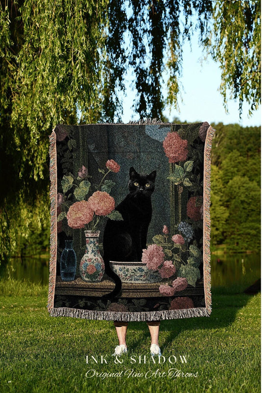 May include: A tapestry throw blanket featuring a black cat sitting amidst floral arrangements in a window. The throw has a dark background with pink and blue flowers, vases, and a decorative border. The text "INK & SHADOW Original Fine Art Throws" is at the bottom.