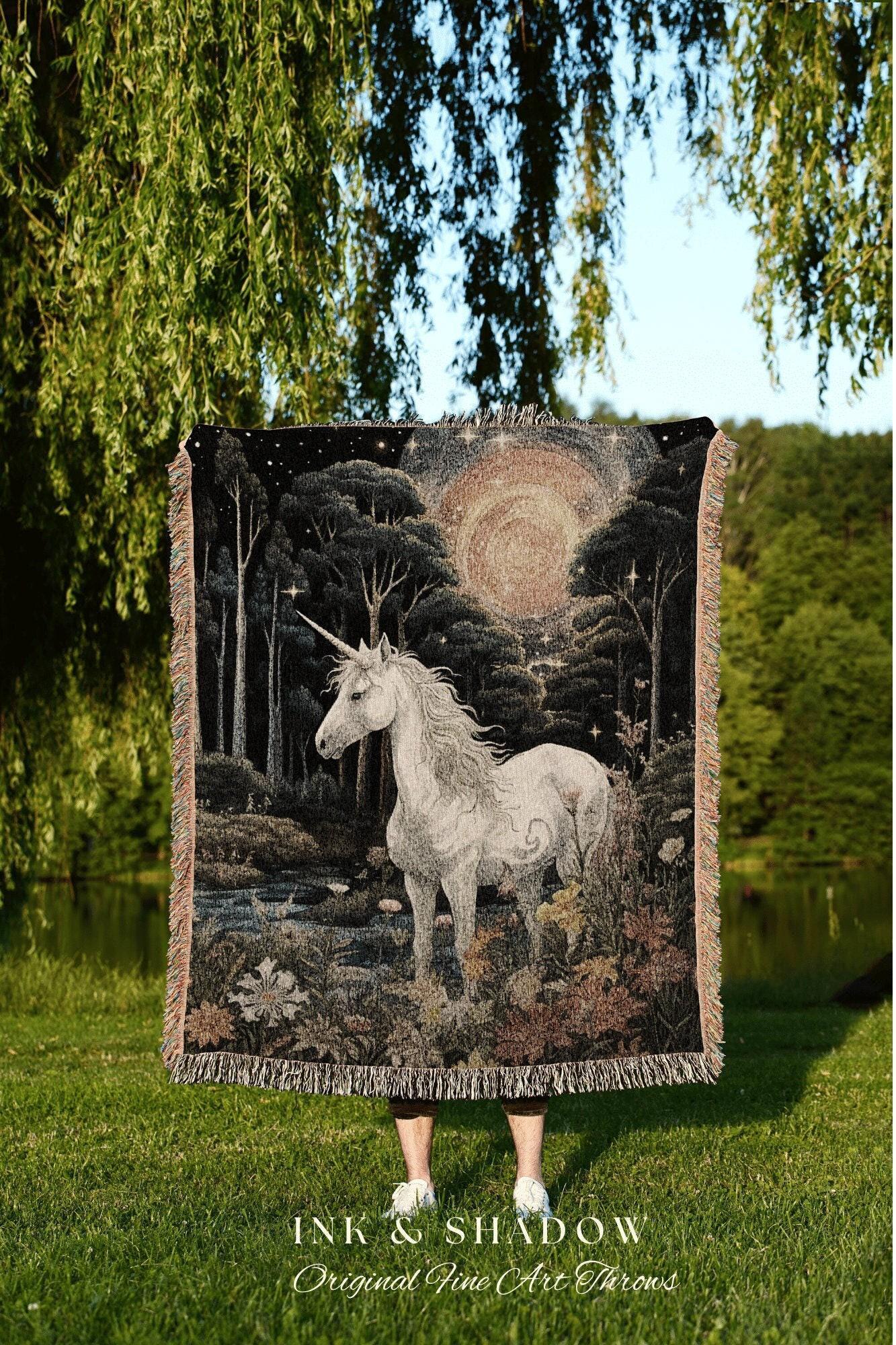 May include: A woven throw blanket featuring a white unicorn in a forest scene with a starry night sky and a large sun. The blanket has a fringed edge and the text "INK & SHADOW Original Fine Art Throws".