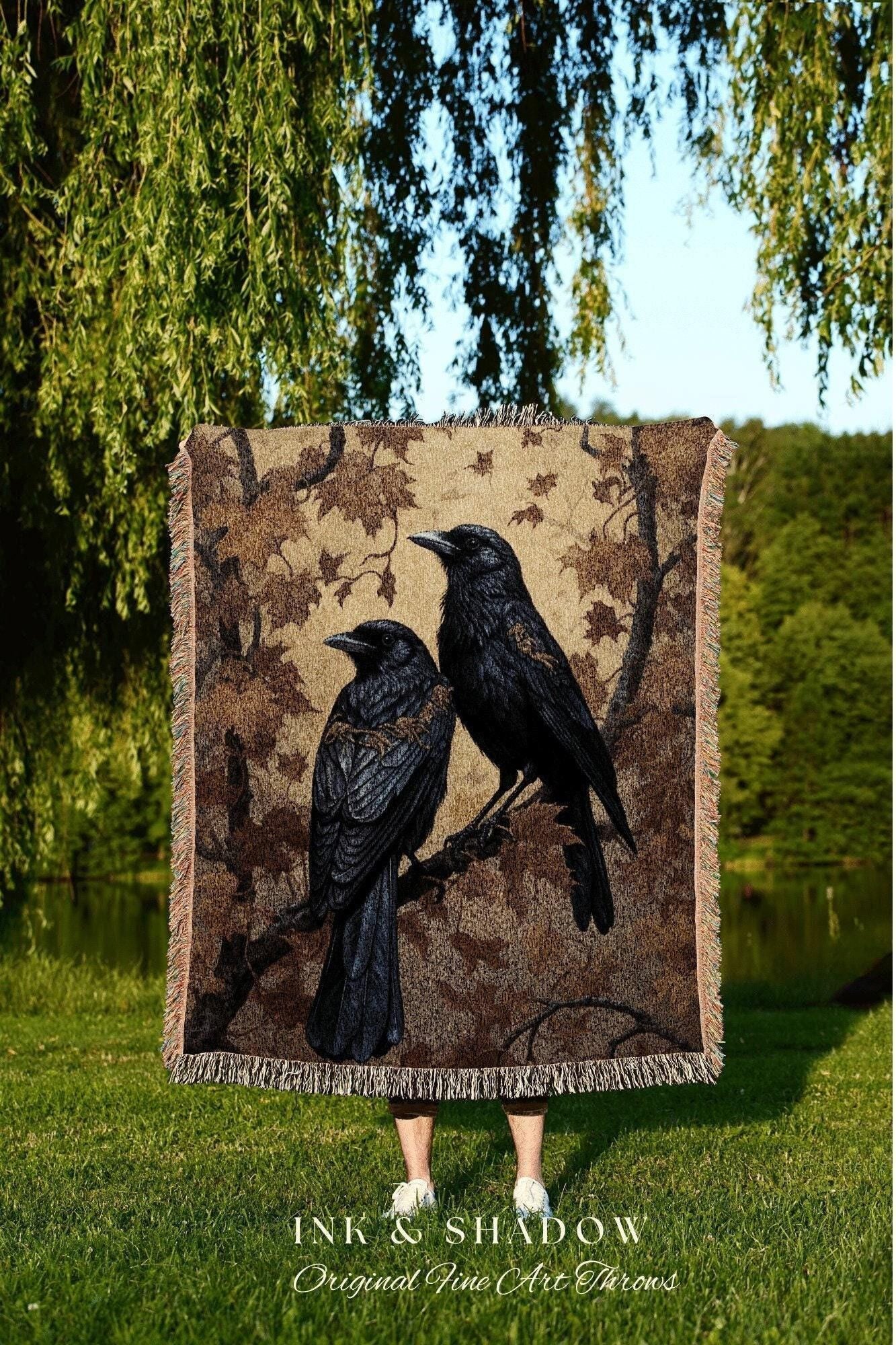May include: A woven throw blanket featuring two black crows perched on a branch, set against a brown and tan background with a leaf pattern. The blanket has a fringed edge and the text "INK & SHADOW Original Fine Art Throws".