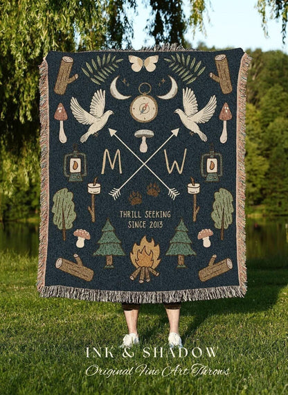 May include: Dark blue throw blanket with a nature-themed design. Features illustrations of doves, arrows, trees, mushrooms, and a campfire. Text includes "M W" and "Thrill Seeking Since 2013". The blanket has a fringed edge.