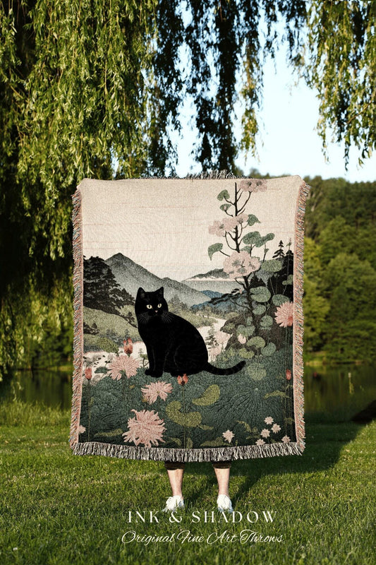 May include: A woven throw blanket featuring a black cat sitting in a landscape with mountains, flowers, and trees. The blanket has a fringed edge and the text "INK & SHADOW Original Fine Art Throws" at the bottom.