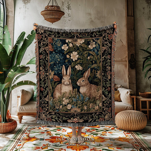 May include: A woven tapestry throw blanket featuring two rabbits amidst a floral and foliage design. The blanket has a black border with a fringe edge. The text "INK & SHADOW Original Fine Art Throws" is printed at the bottom.