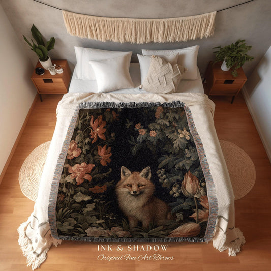 May include: A woven throw blanket featuring a detailed illustration of a fox amidst flowers and foliage. The color palette includes shades of brown, green, and orange against a dark background. The blanket has a fringed edge and the text "INK & SHADOW Original Fine Art Throws".