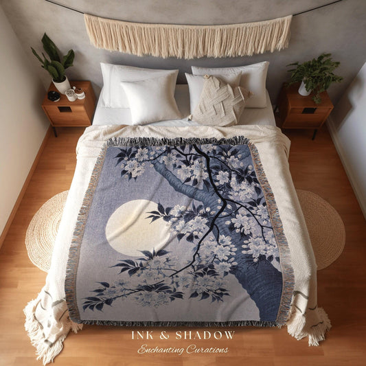 May include: A woven throw blanket featuring a tree with white blossoms and a full moon against a blue and gray background. The blanket has a fringed edge and the text "INK & SHADOW Enchanting Curations".