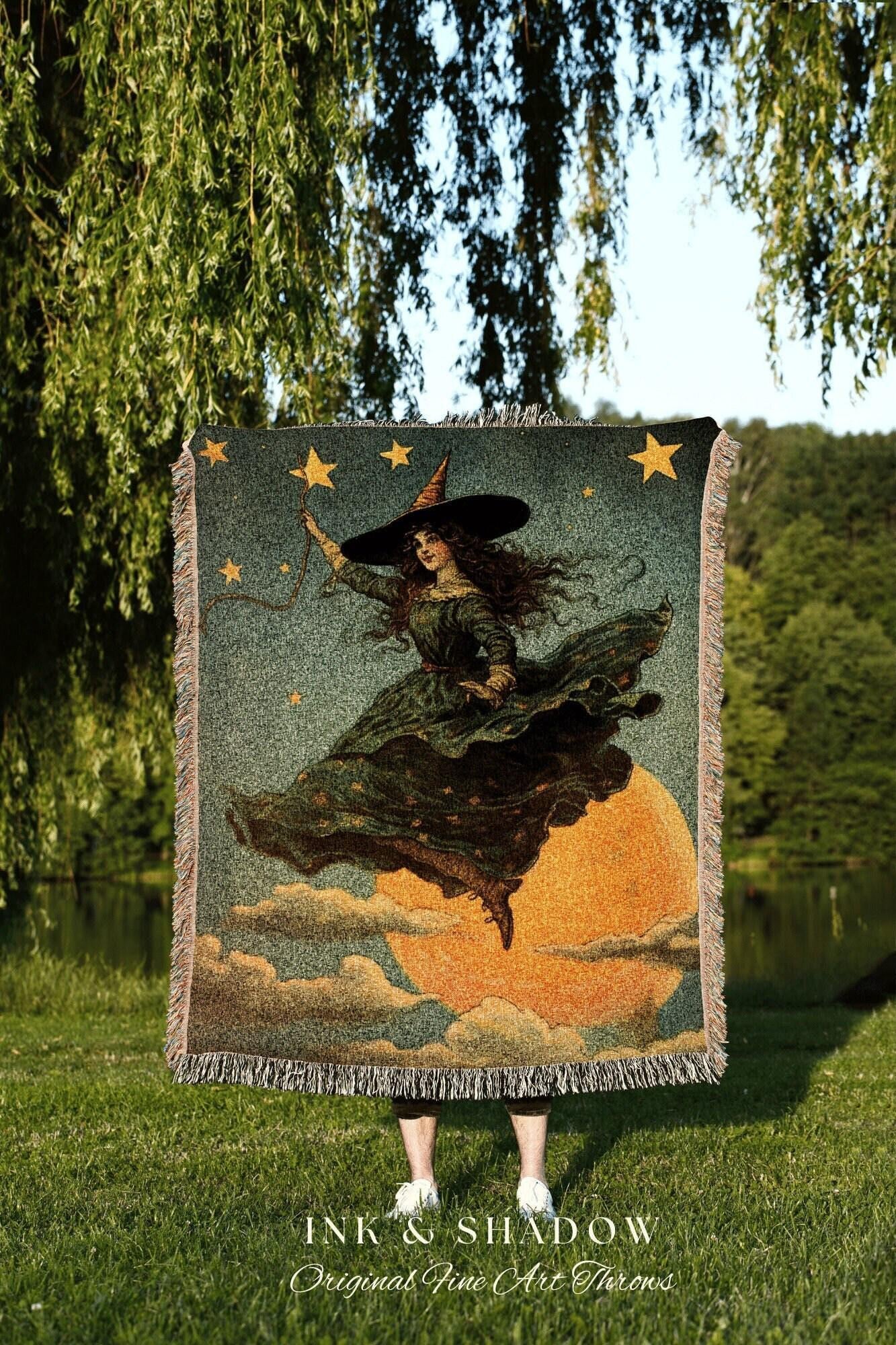 May include: A tapestry throw blanket featuring a vintage illustration of a witch flying on a broomstick in front of a full moon. The witch wears a black hat and green dress. The blanket has a fringe border and the text "INK & SHADOW Original Fine Art Throws".