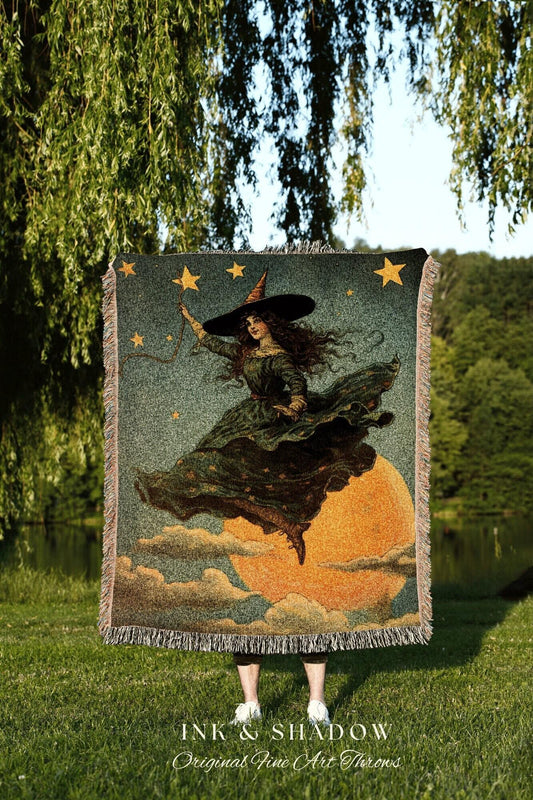 May include: A tapestry throw blanket featuring a vintage illustration of a witch flying on a broomstick in front of a full moon. The witch wears a black hat and green dress. The blanket has a fringe border and the text "INK & SHADOW Original Fine Art Throws".