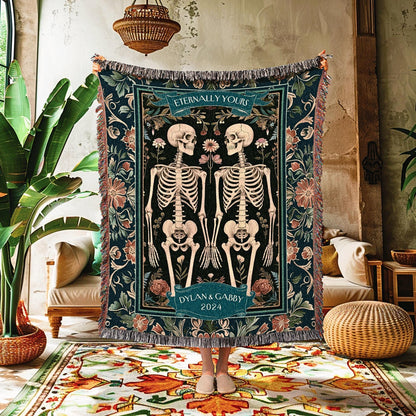 May include: A dark teal woven throw blanket featuring a design of two skeletons holding hands, surrounded by floral patterns. The blanket has the text "Eternally Yours" and "Dylan & Gabby 2024". The edges are fringed.