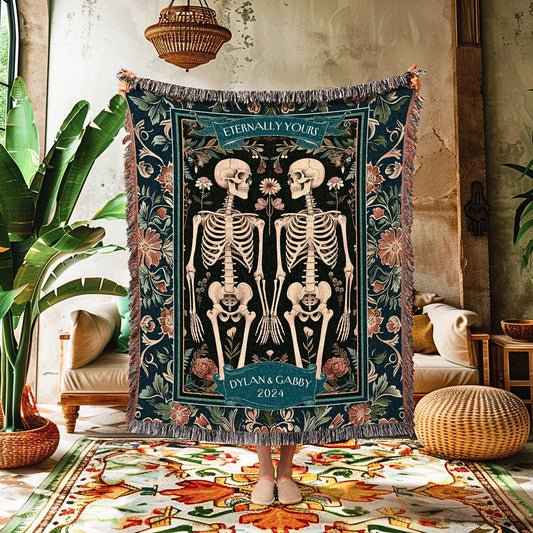 May include: A dark teal woven throw blanket featuring a design of two skeletons holding hands, surrounded by floral patterns. The blanket has the text "Eternally Yours" and "Dylan & Gabby 2024". The edges are fringed.