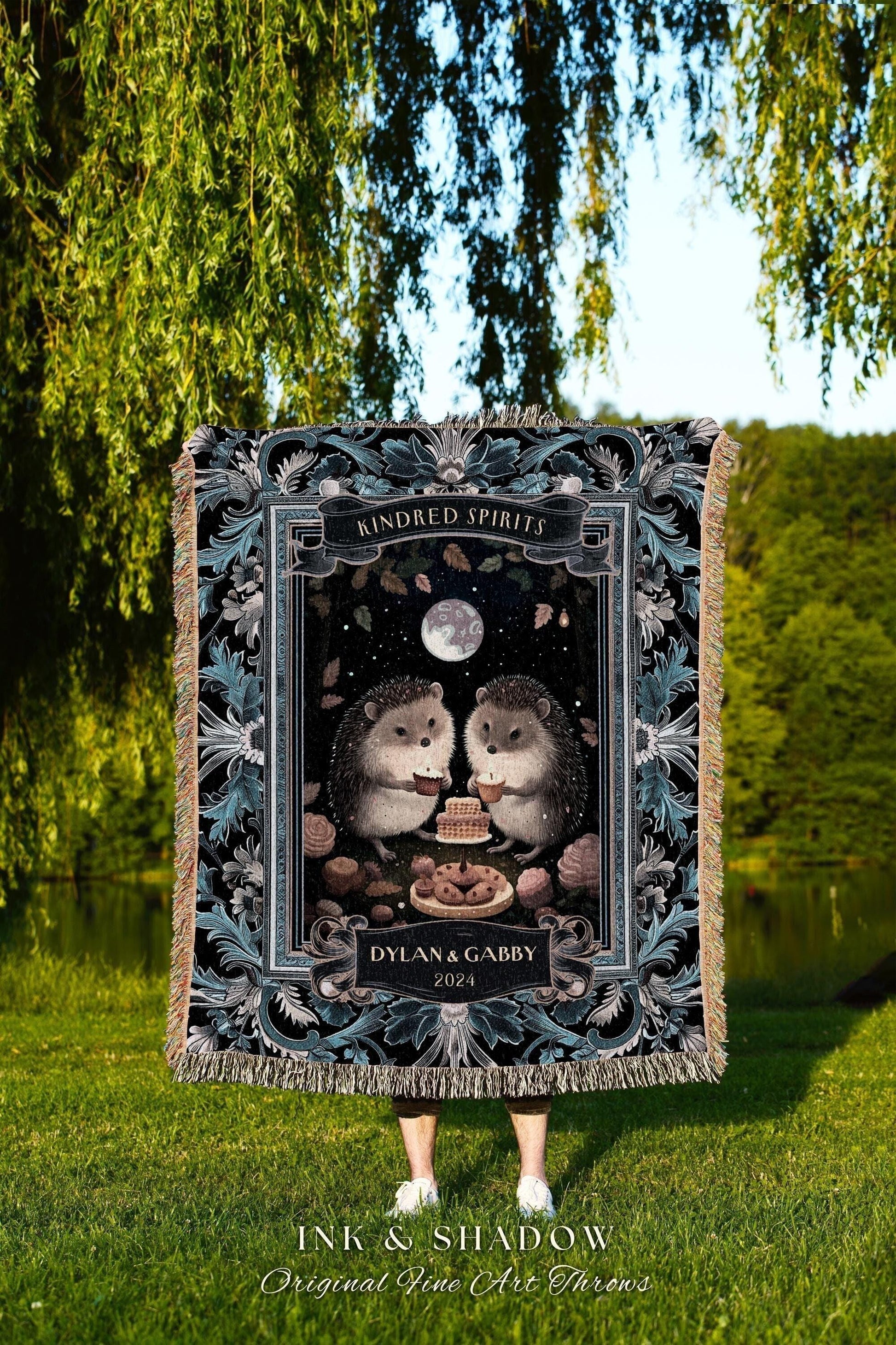 May include: A decorative throw blanket featuring a whimsical illustration of two hedgehogs sharing a cake under a full moon. The blanket has a black background with a decorative border and the text "Kindred Spirits" and "Dylan & Gabby 2024".