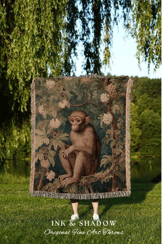 May include: A tapestry throw blanket featuring a detailed illustration of a monkey sitting in a tree, surrounded by foliage and flowers. The blanket has a fringed edge and the text "INK & SHADOW Original Fine Art Throws" at the bottom.