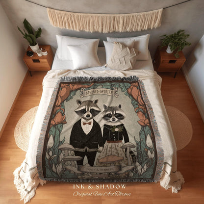 May include: A cream-colored throw blanket featuring a whimsical illustration of two raccoons dressed in formal attire, framed by floral and mushroom motifs. The text "Kindred Spirits" and "INK & SHADOW Original Fine Art Throws" are visible. The blanket is draped over a bed.