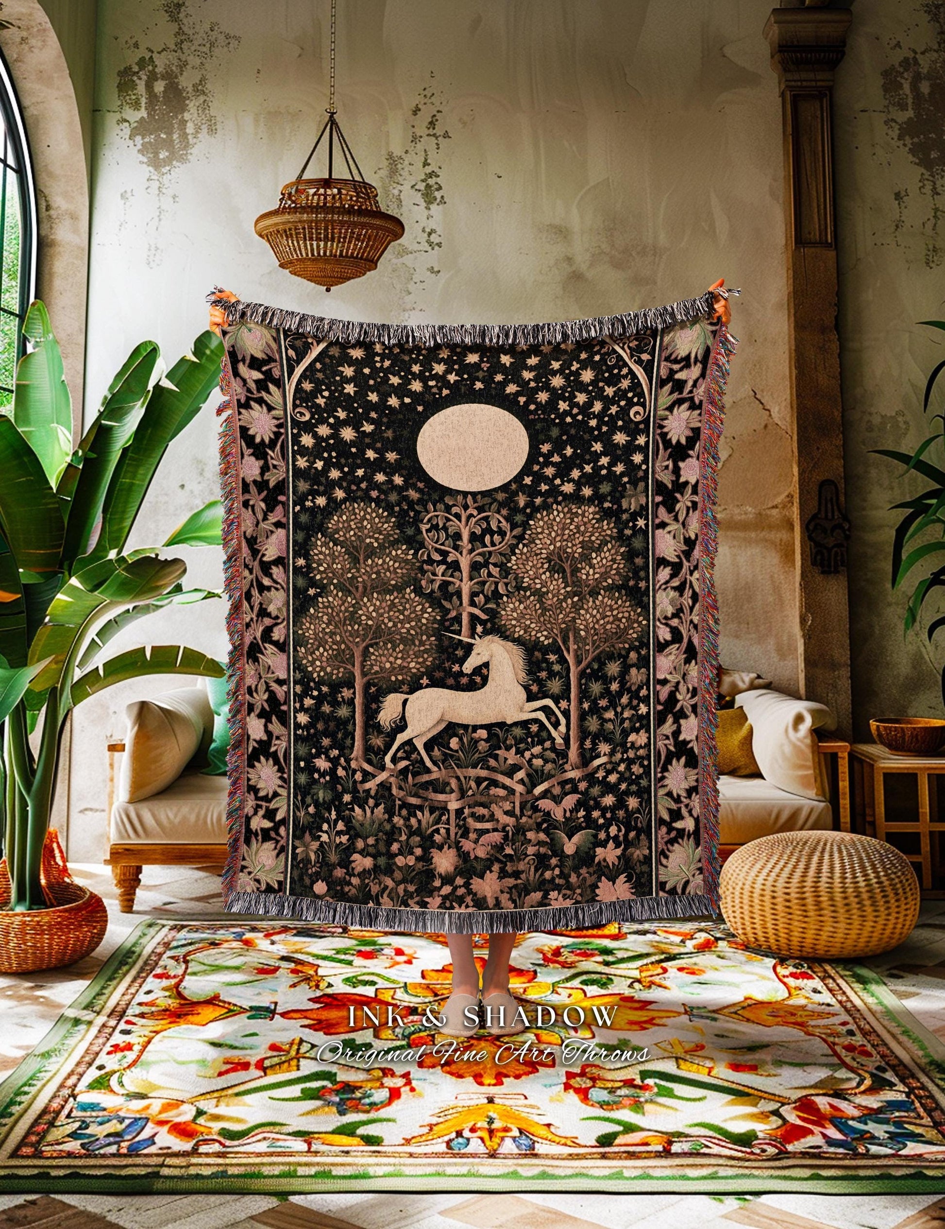 May include: A tapestry featuring a white unicorn in a forest scene with a full moon and stars. The tapestry has a black background with a fringed edge and is held up in front of a colorful rug. The text "INK & SHADOW Original Fine Art Throws" is visible.