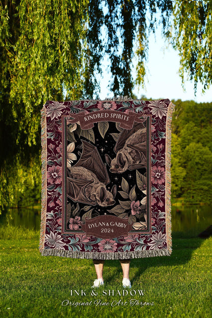 May include: A decorative throw blanket featuring a dark background with illustrations of bats, flowers, and stars. The blanket has a burgundy border with floral patterns and the text "KINDRED SPIRITS" and "DYLAN & GABBY 2024". The blanket is fringed.