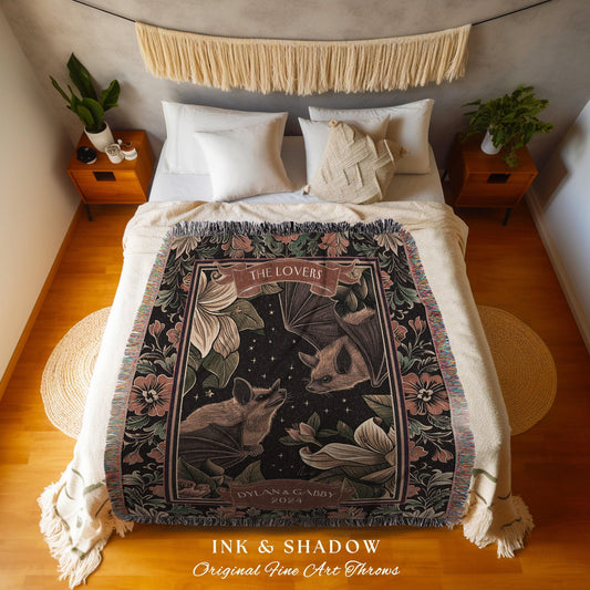 May include: A decorative throw blanket featuring a design with two bats, flowers, and the text "THE LOVERS". The blanket has a black background with pink, green, and brown accents. The text "INK & SHADOW" and "Original Fine Art Throws" is also visible.