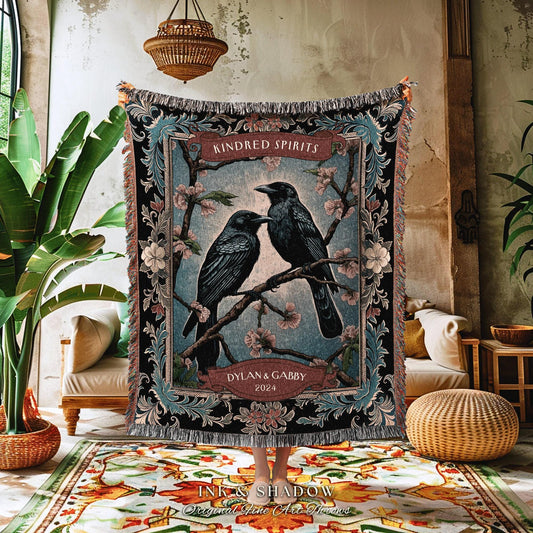 May include: A woven throw blanket featuring two black ravens perched on a flowering branch, with the text "Kindred Spirits" and "Dylana & Gabby 2024". The blanket has a black border with floral and scroll details, and fringed edges.
