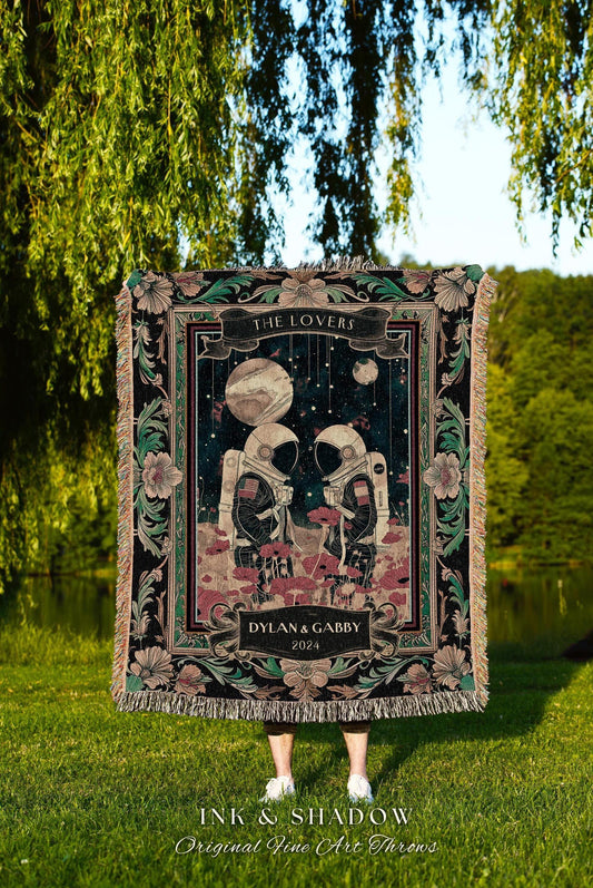 May include: A black throw blanket with a floral border and fringed edges. The center features an illustration of two astronauts under a starry sky, with the text "THE LOVERS" and "DYLAN & GABBY 2024". The blanket is held outdoors.