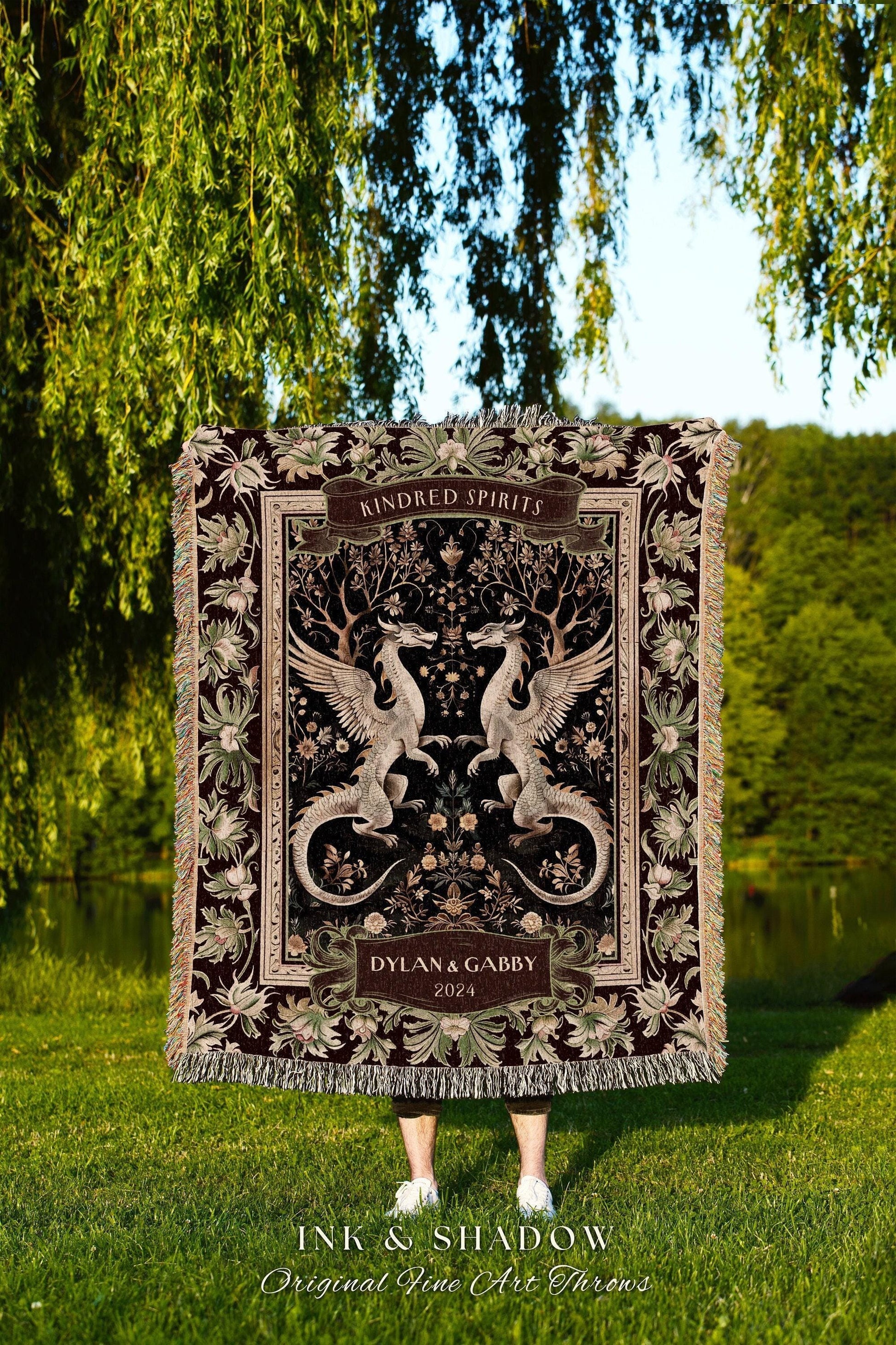 May include: A burgundy throw blanket with a detailed design featuring two winged dragons facing each other. The blanket has a floral border and the words "Kindred Spirits" and "Dylan & Gabby 2024". The blanket is fringed and has the text "INK & SHADOW Original Fine Art Throws".