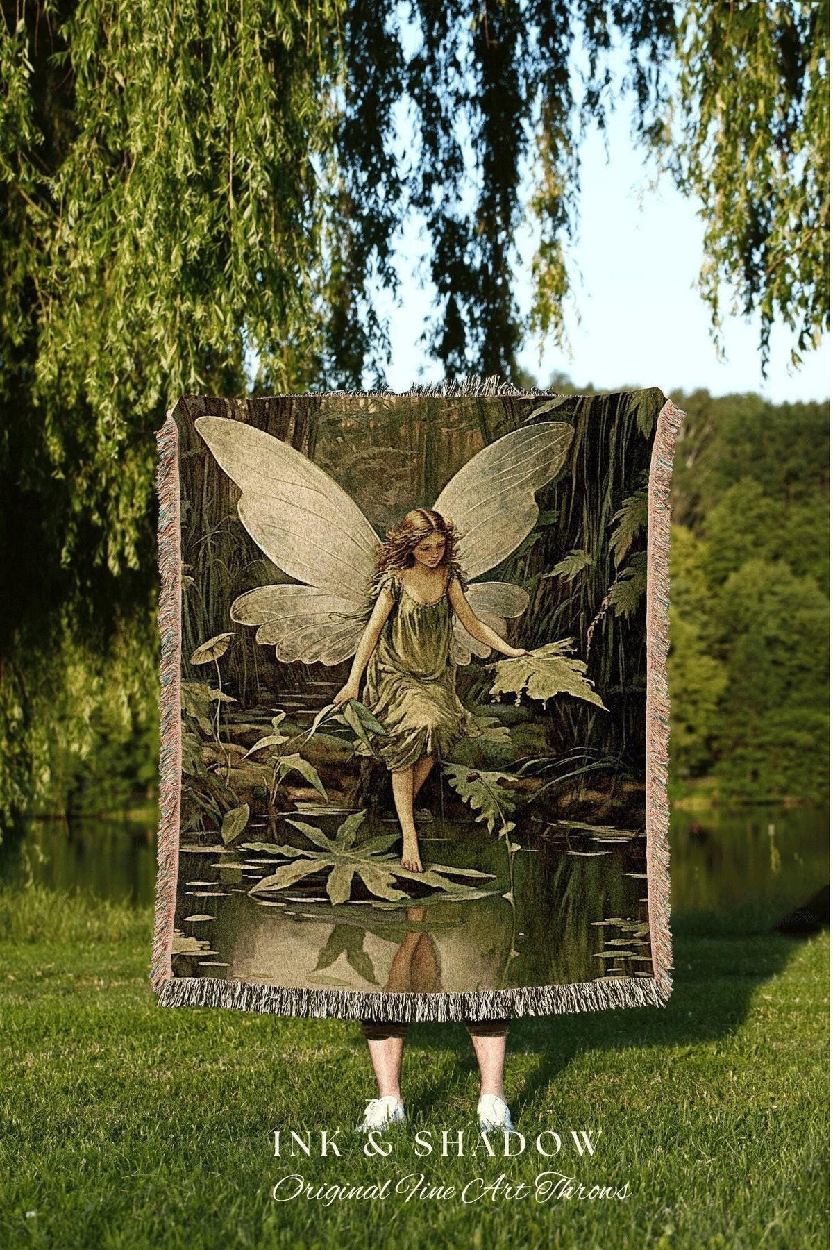May include: A woven throw blanket featuring a fairy in a forest scene with a pond. The fairy has large wings and is wearing a green dress. The blanket has a fringed edge and the text "INK & SHADOW Original Fine Art Throws".