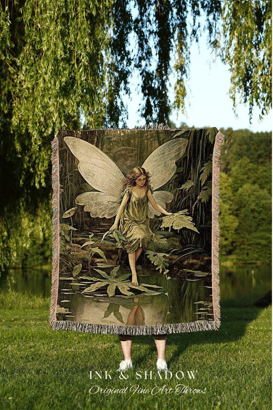May include: A woven throw blanket featuring a fairy in a forest scene with a pond. The fairy has large wings and is wearing a green dress. The blanket has a fringed edge and the text "INK & SHADOW Original Fine Art Throws".