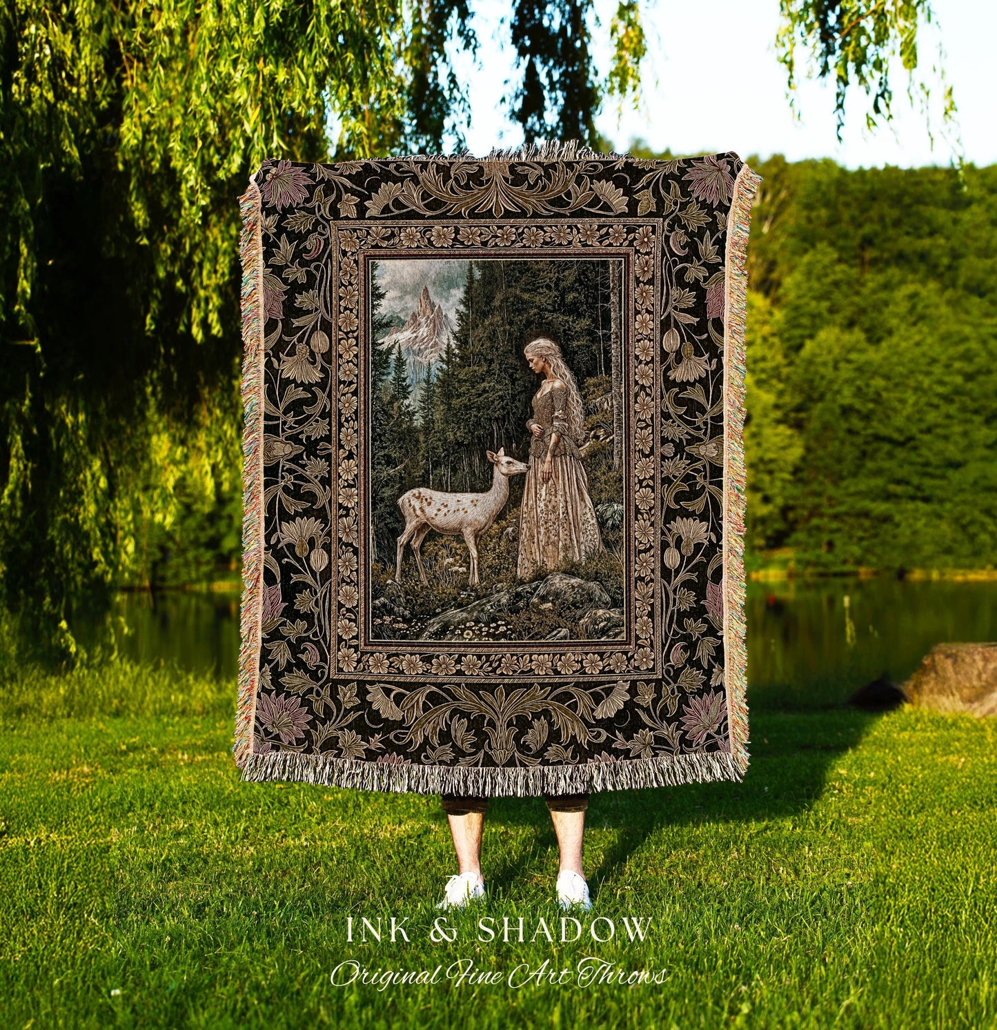 May include: A decorative throw blanket featuring a detailed forest scene with a woman and a deer. The blanket has a black background with floral and leaf border designs and fringe. The text "INK & SHADOW Original Fine Art Throws" is at the bottom.