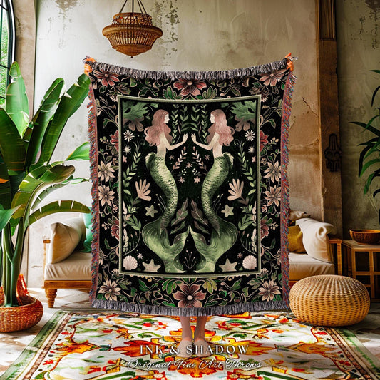 May include: A black throw blanket featuring two mermaids with pink hair and green tails, surrounded by floral and botanical designs. The blanket has a fringed edge and the text "INK & SHADOW Original Fine Art Throws".