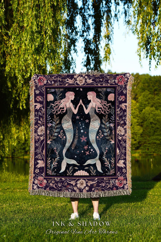 May include: A dark purple throw blanket featuring two mermaids with flowing hair, facing each other. The blanket has a floral and seashell border, with the text "INK & SHADOW Original Fine Art Throws" at the bottom. The blanket is fringed.
