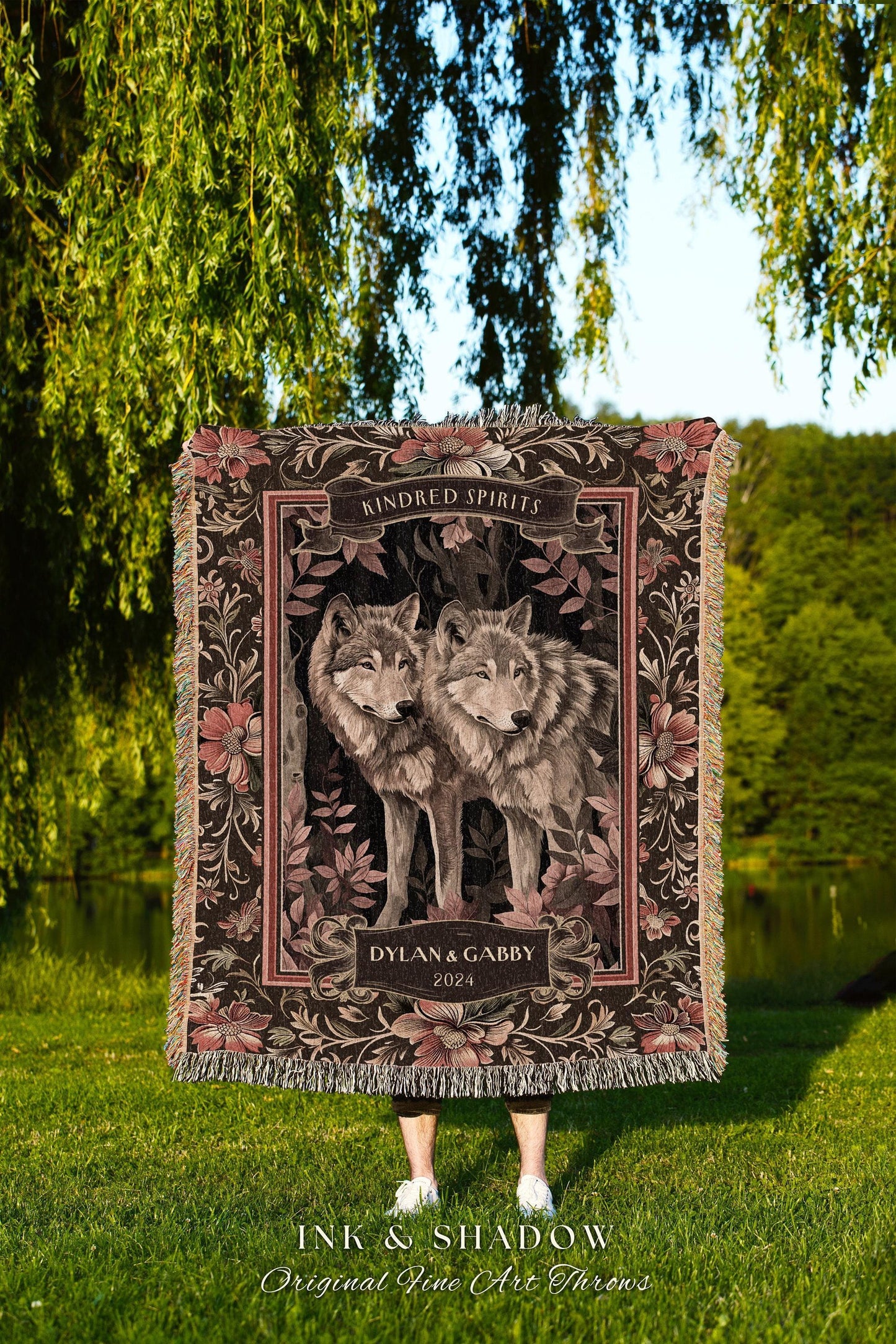 May include: A woven throw blanket featuring two wolves, surrounded by floral patterns in shades of brown and pink. The words "Kindred Spirits" and "Dylan & Gabby 2024" are displayed on the blanket. The blanket has a fringed edge.