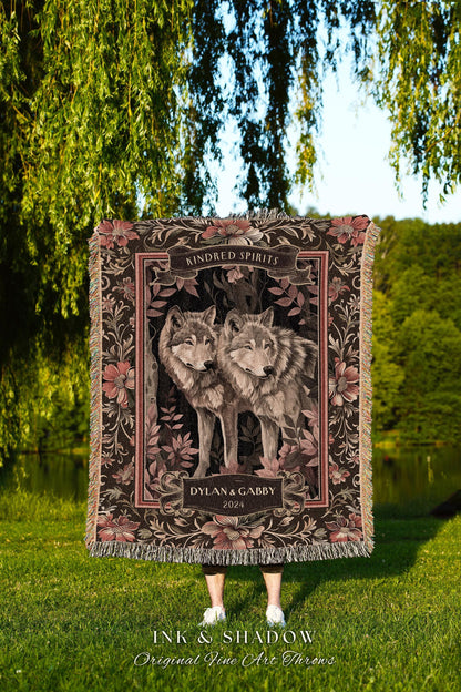 May include: A woven throw blanket featuring two wolves, surrounded by floral patterns in shades of brown and pink. The words "Kindred Spirits" and "Dylan & Gabby 2024" are displayed on the blanket. The blanket has a fringed edge.