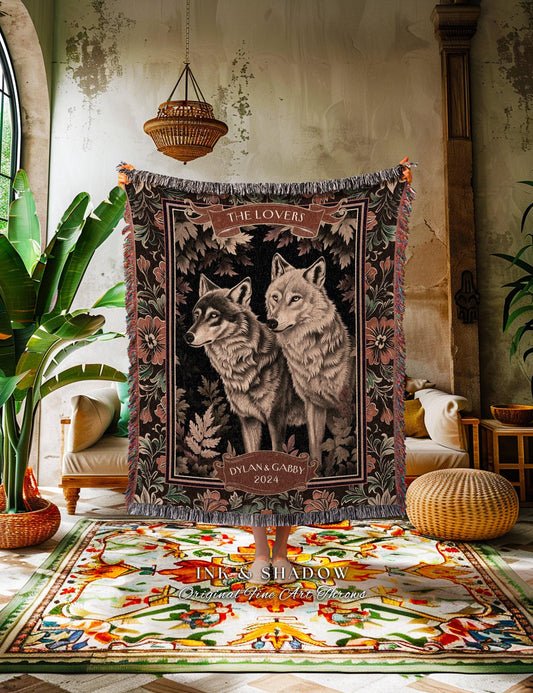 May include: A woven tapestry blanket featuring two wolves, with the text "THE LOVERS" and "DYLAN & GABBY 2024". The blanket has a dark background with floral and leaf patterns, and fringed edges. The wolves are depicted in shades of gray and brown.
