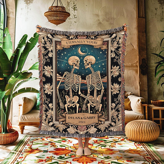 May include: A woven tapestry blanket featuring a romantic scene with two skeletons facing each other under a starry night sky. The blanket has a dark blue background with the words "Eternally Yours" and "Dylan & Gabby 2024". The edges are fringed.