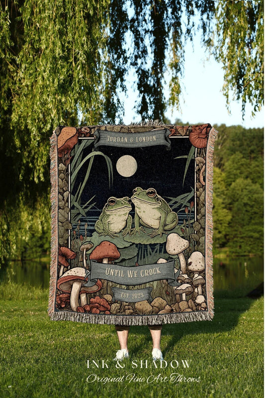 May include: A woven throw blanket featuring a whimsical illustration of two frogs on lily pads under a full moon. The design includes mushrooms and the text "UNTIL WE CROCK EST 2023" and "JORDAN & LONDON". The blanket has a fringed edge.