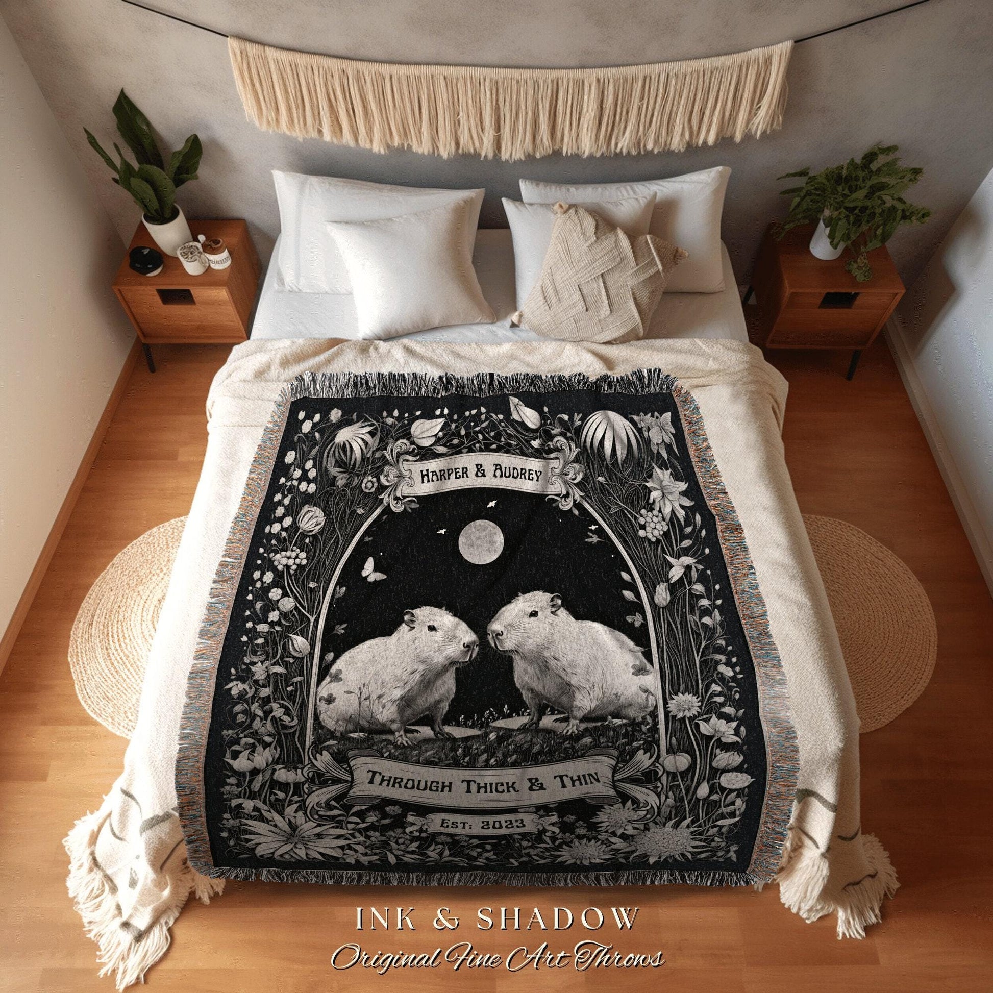 May include: A black and white throw blanket featuring two capybaras under a full moon, surrounded by floral and botanical illustrations. The blanket has the text "Harper & Audrey" and "Through Thick & Thin, Est. 2023". The blanket is on a bed with white pillows.