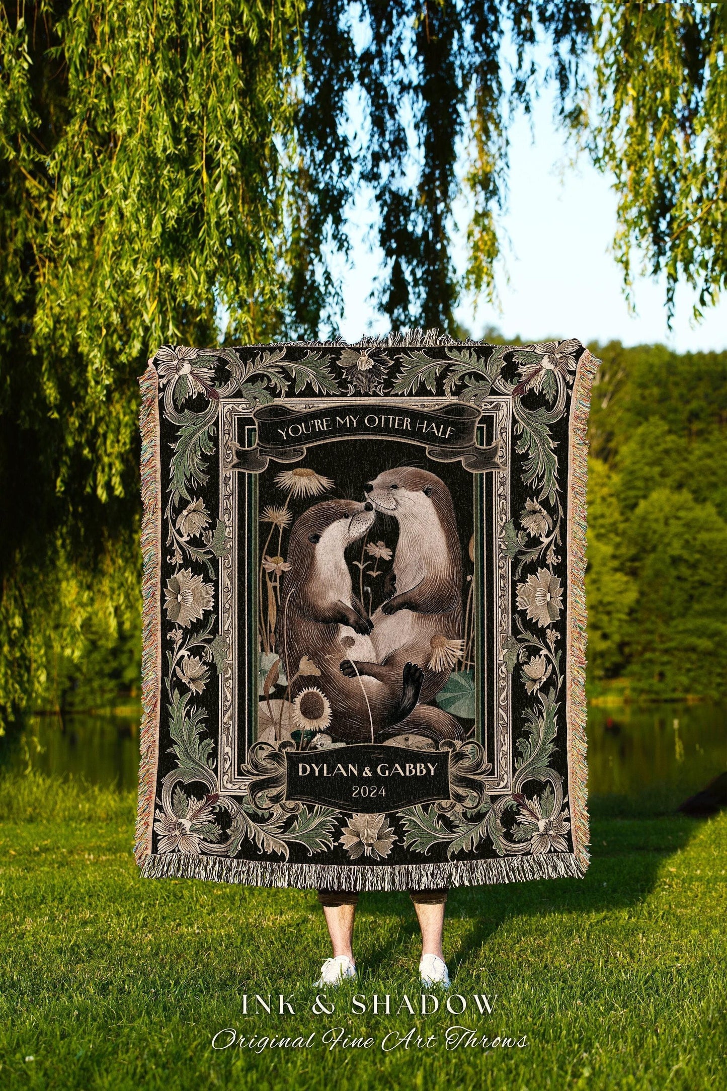 May include: A black throw blanket with a decorative border and fringe. The center features an illustration of two otters with the text "YOU'RE MY OTTER HALF" and "DYLAN & GABBY 2024". The blanket is displayed outdoors.