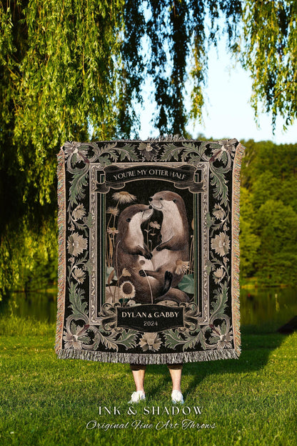 May include: A black throw blanket with a decorative border and fringe. The center features an illustration of two otters with the text "YOU'RE MY OTTER HALF" and "DYLAN & GABBY 2024". The blanket is displayed outdoors.
