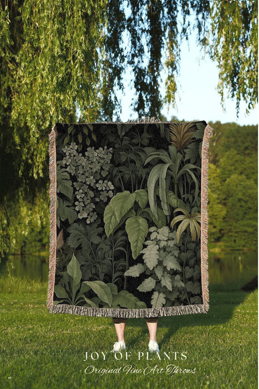 May include: A dark green throw blanket featuring a botanical print of various plants and foliage. The blanket has a fringed edge and the text "JOY OF PLANTS Original Fine Art Throws" at the bottom. The blanket is held up in a grassy area.